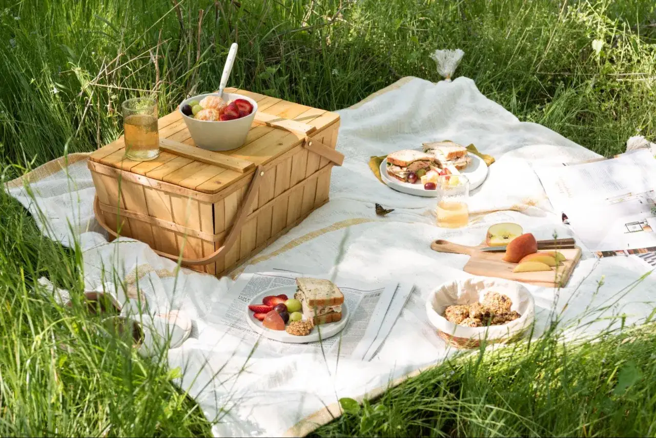 Picknickdecke mit Picknickkorb, Sandwiches, Obst und Keksen im Gras. Ein Schmetterling sitzt auf der Decke.