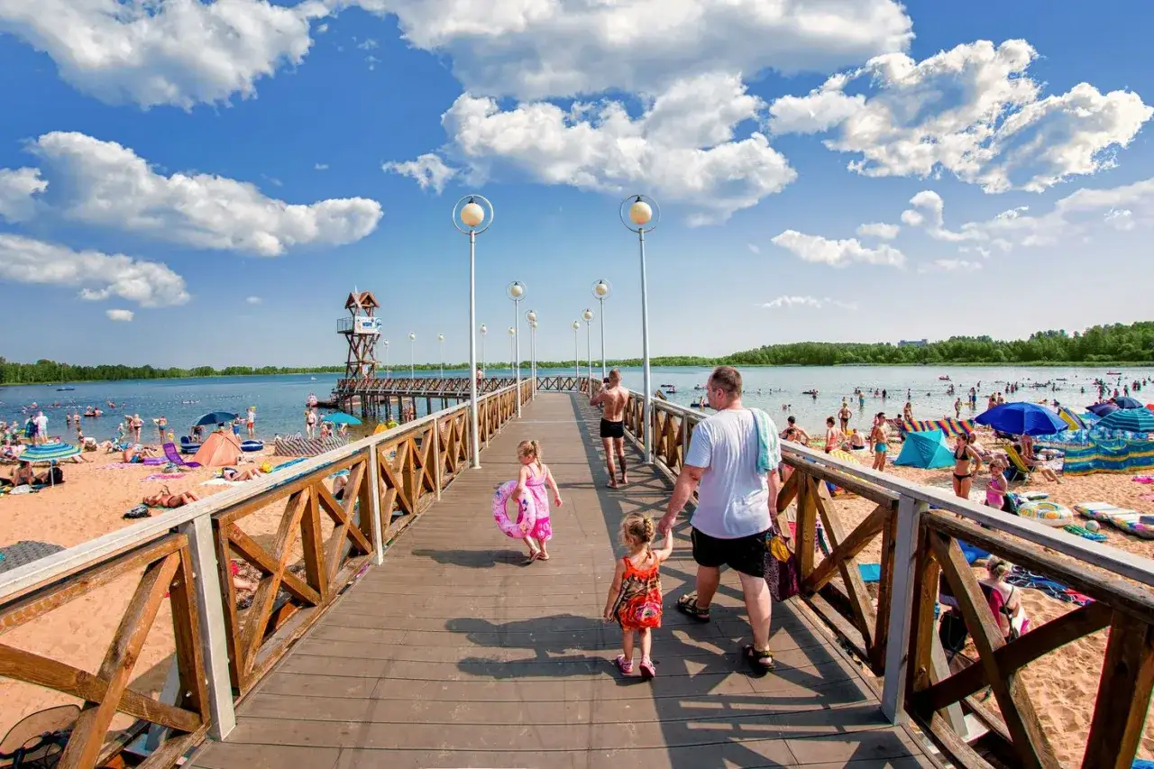 Rodzina spaceruje po molo nad jeziorem Pogoria 3. Słoneczny dzień, plaża pełna ludzi, woda zachęca do kąpieli.