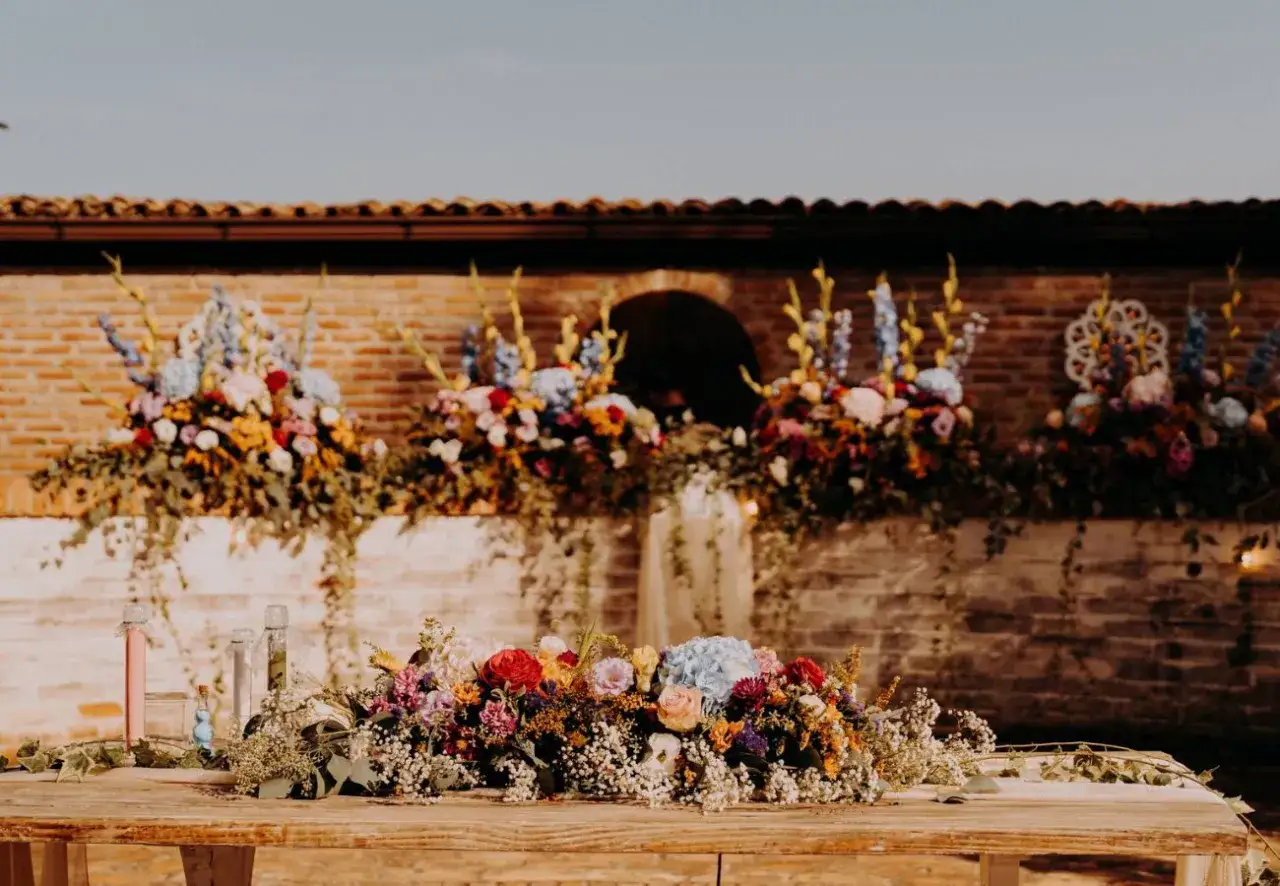 Un tavolo in legno addobbato con fiori colorati, perfetto per decidere quando sposarsi. Dietro, composizioni floreali pendenti su un muro di mattoni.