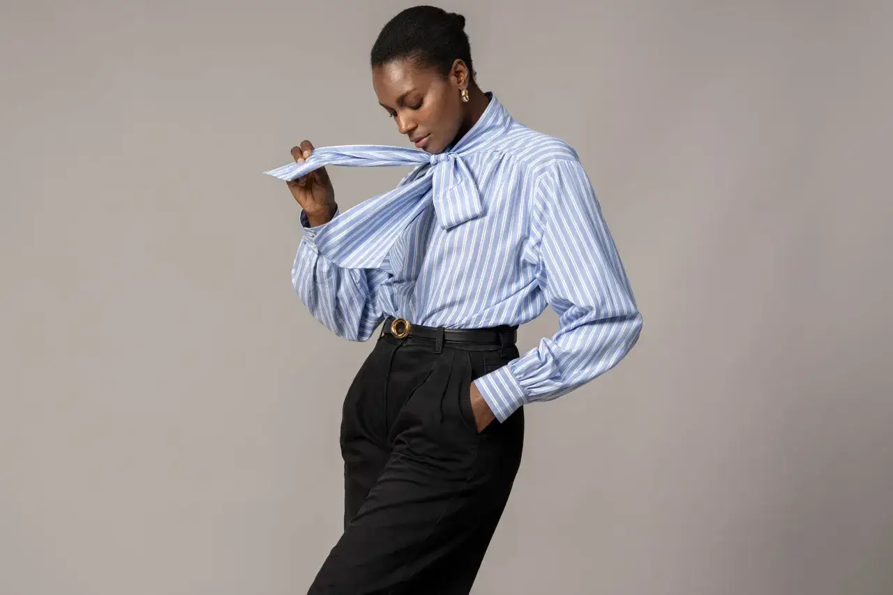Une femme porte un **outfit mariniere** chic : chemise rayée bleu et blanc à nœud, pantalon noir taille haute.