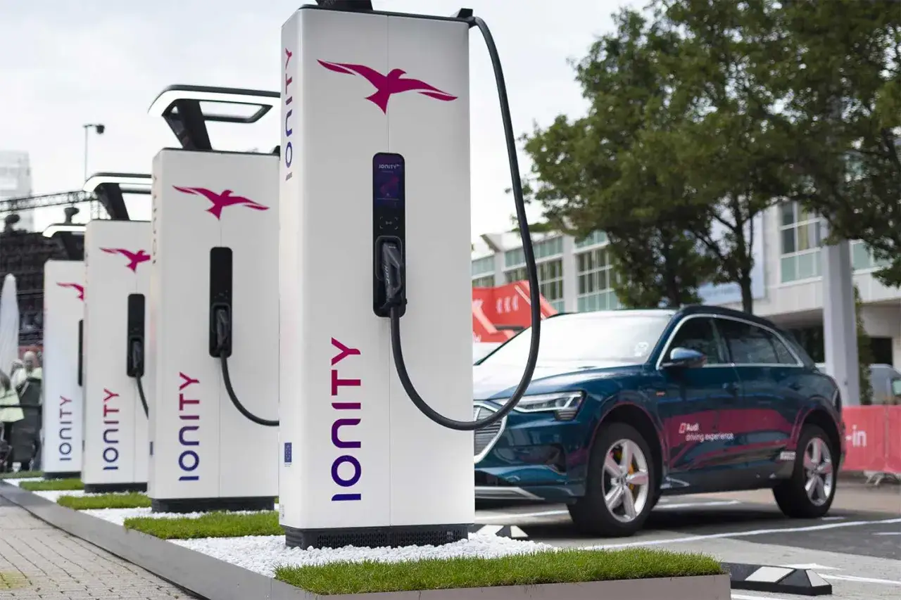 Plusieurs bornes de recharge IONITY, avec leur logo d'oiseau rose, sont align&eacute;es. Une voiture &eacute;lectrique bleue est gar&eacute;e &agrave; c&ocirc;t&eacute; de l'une d'elles, pr&ecirc;te &agrave; &ecirc;tre recharg&eacute;e.