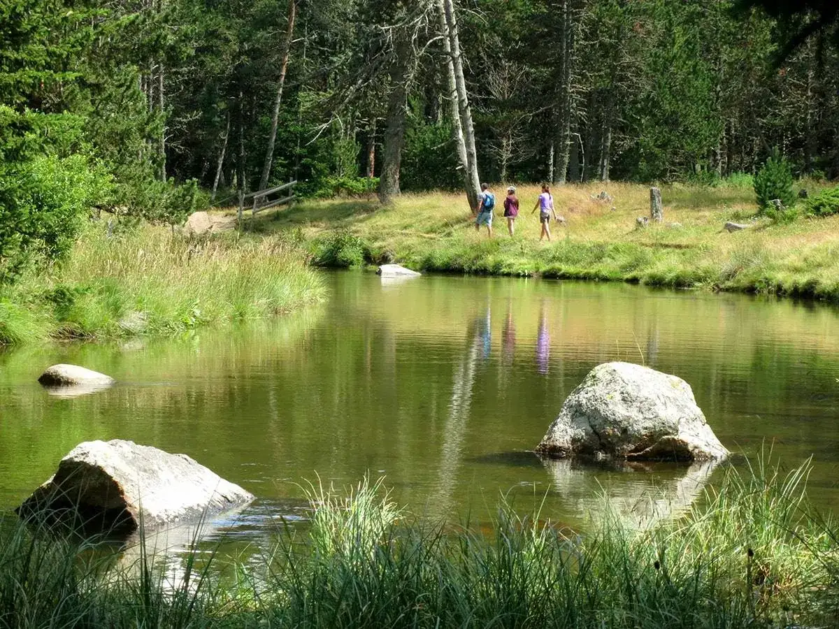 Un estany de l'Orri ser&egrave; amb roques i tres excursionistes a la vora, envoltat de pins i vegetaci&oacute;.