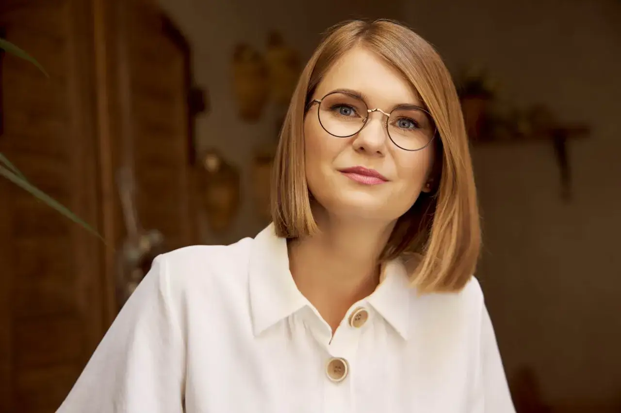 Kobieta w białej koszuli z guzikami, nosząca okulary w cienkich oprawkach, patrzy wprost przed siebie.
