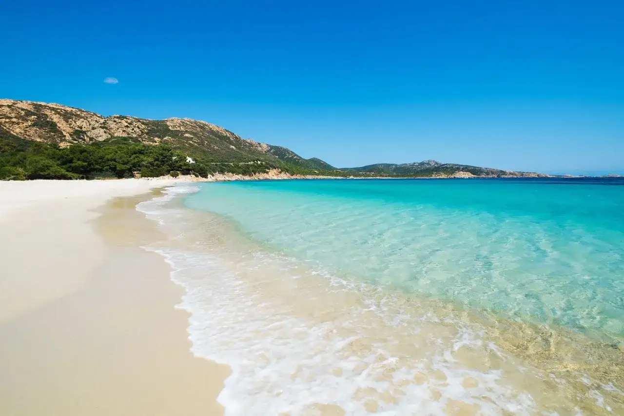 Plage de sable blanc bordée par une eau turquoise cristalline, avec des collines verdoyantes en arrière-plan. Un coin de paradis sur la **riviera italienne**.