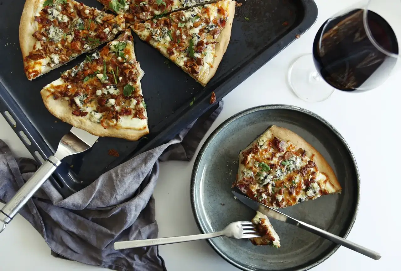 Pyszna pizza z kurkami, serem pleśniowym i boczkiem na blasze, obok talerz z kawałkiem i kieliszek czerwonego wina.