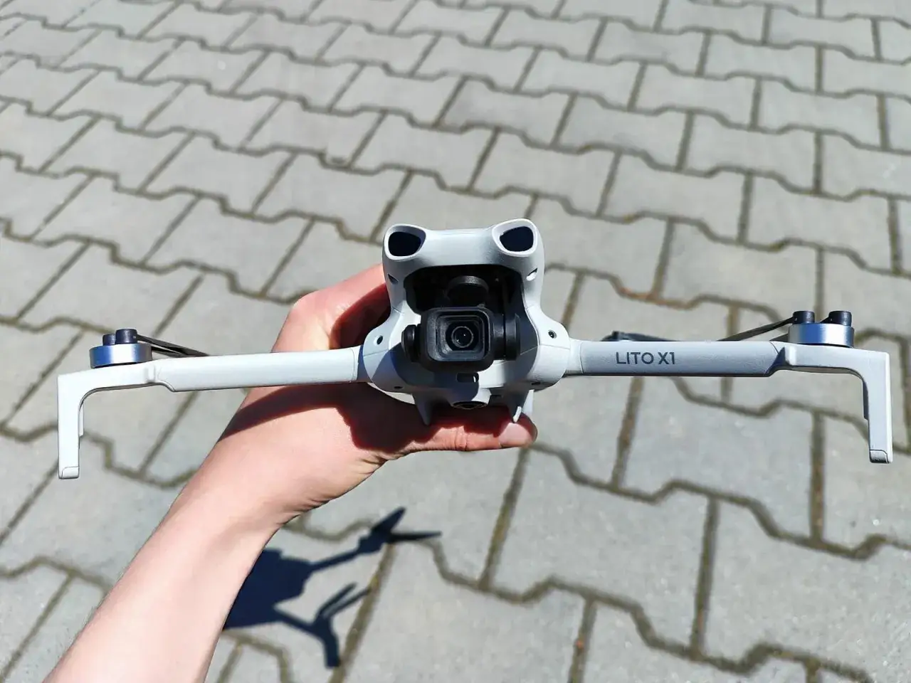 Biały dron DJI LITO X1 z kamerą, trzymany w dłoni na tle chodnika.