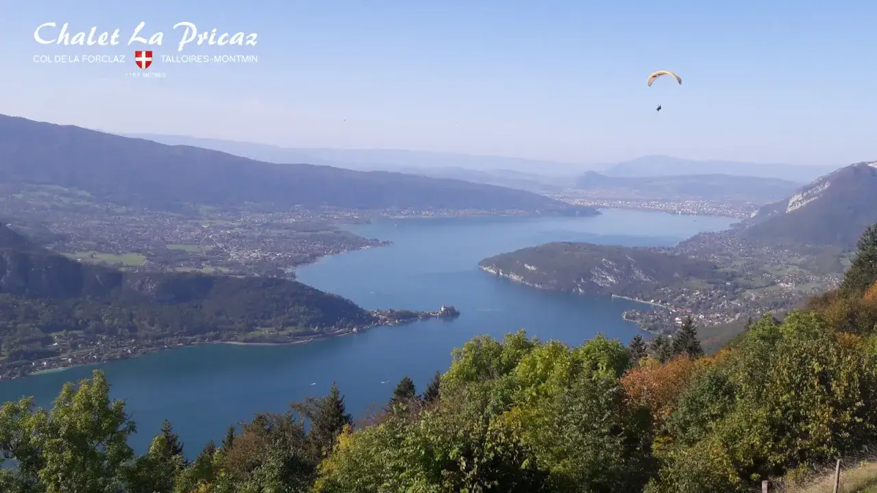 Vue panoramique du lac d'Annecy depuis le Col de la Forclaz. Un parapentiste survole le lac, offrant une perspective aérienne unique.