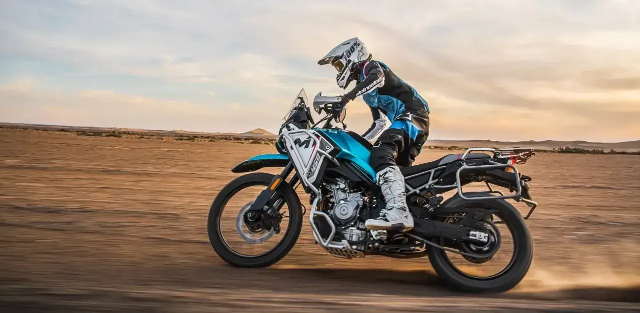 Motocyklista w kasku i stroju terenowym pędzi na turkusowym motocyklu po pustyni. Czy warto kupić CF Moto? Ten sprzęt wygląda na gotowy na każde wyzwanie.