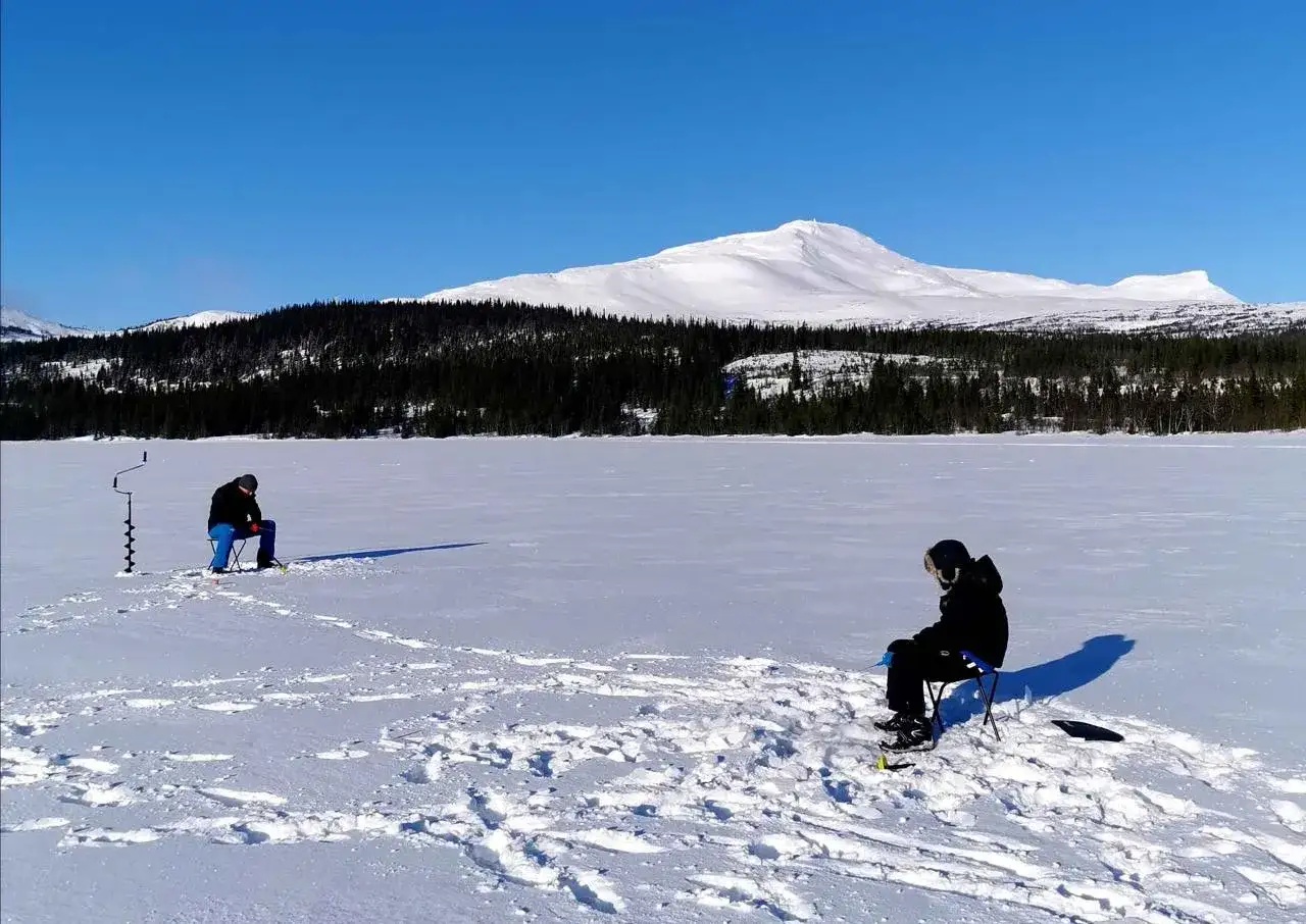 Zwei Angler warten geduldig auf den Fang beim Eisangeln in Schweden. Im Hintergrund erheben sich schneebedeckte Berge und dunkle Tannenwälder.