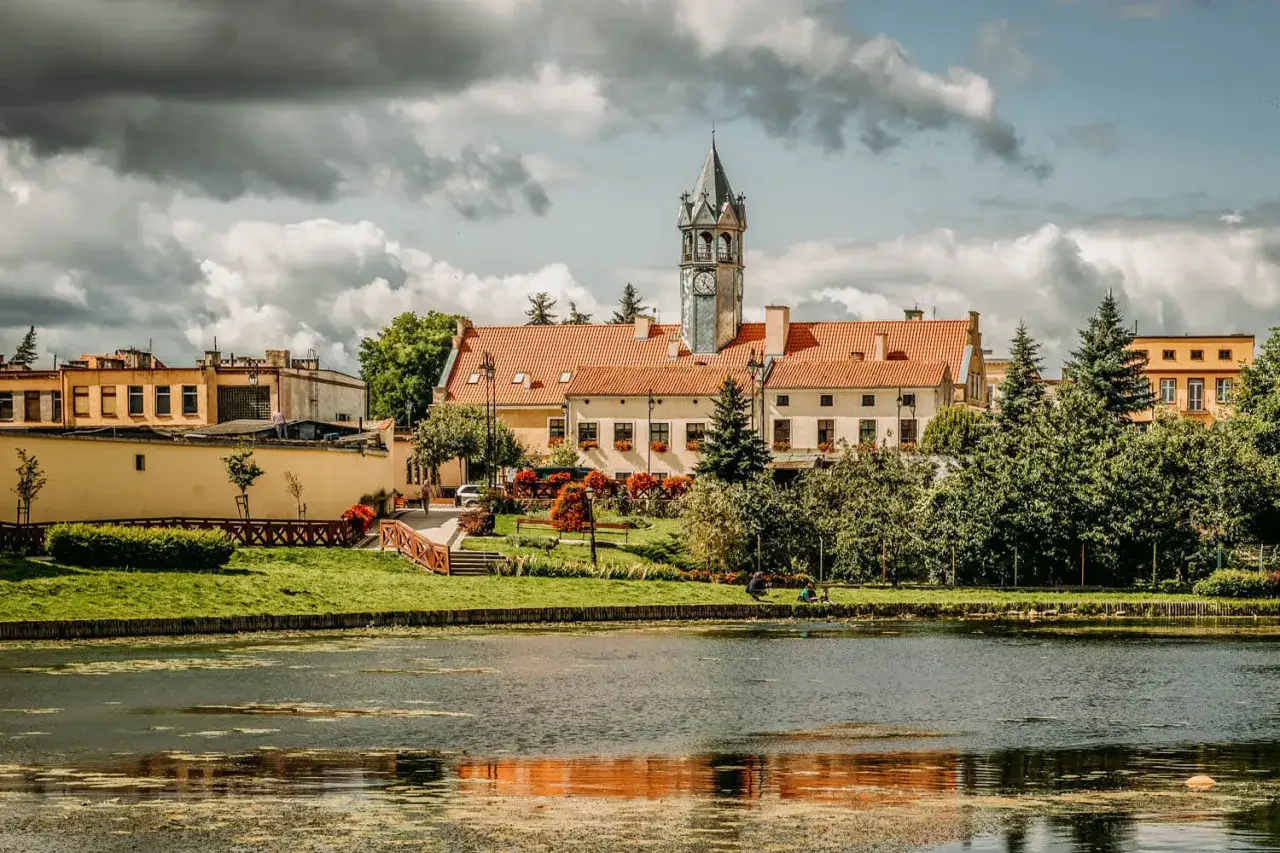 Barczewo atrakcje: malowniczy widok na rynek z wieżą zegarową, kamienice z czerwonymi dachami i zielony park nad jeziorem.