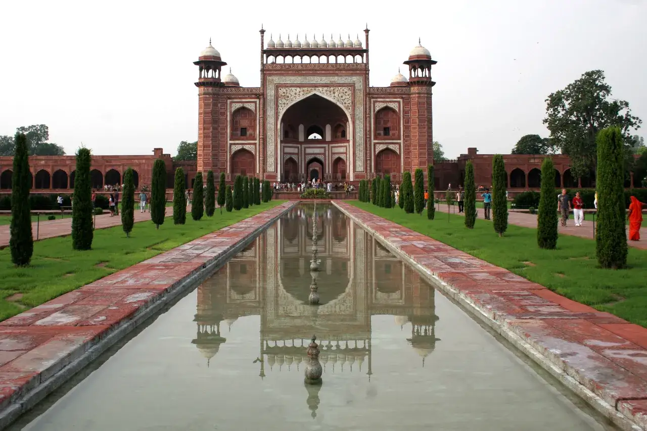 Der Eingang zum Taj Mahal, eine Moschee, spiegelt sich in einem ruhigen Wasserbecken, flankiert von Zypressen und gepflegten Rasenfl&auml;chen.