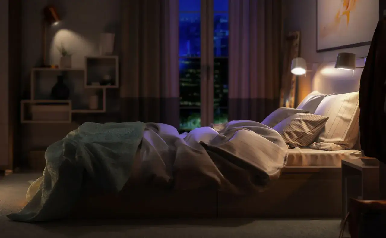 Un letto disfatto con una coperta azzurra, pronto per dormire fuori casa. La luce soffusa illumina la stanza, mentre fuori si intravede la città notturna.