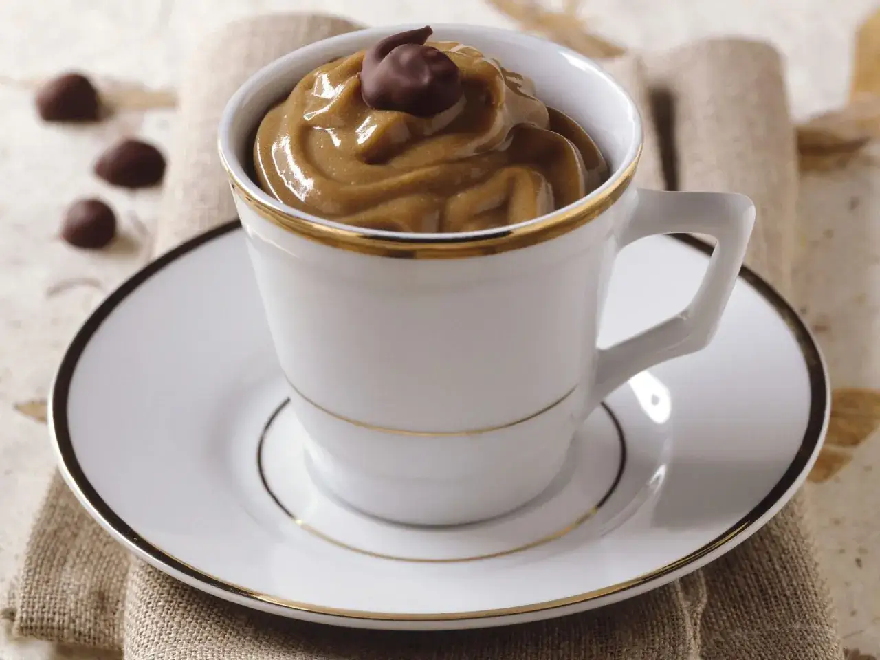Cremige Crema al caffè in einer eleganten Tasse, garniert mit einem Schokoladenstück. Daneben liegen kleine Pralinen.