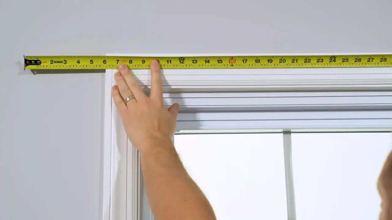 A hand uses a tape measure to determine how to measure curtain length and width across a window frame.