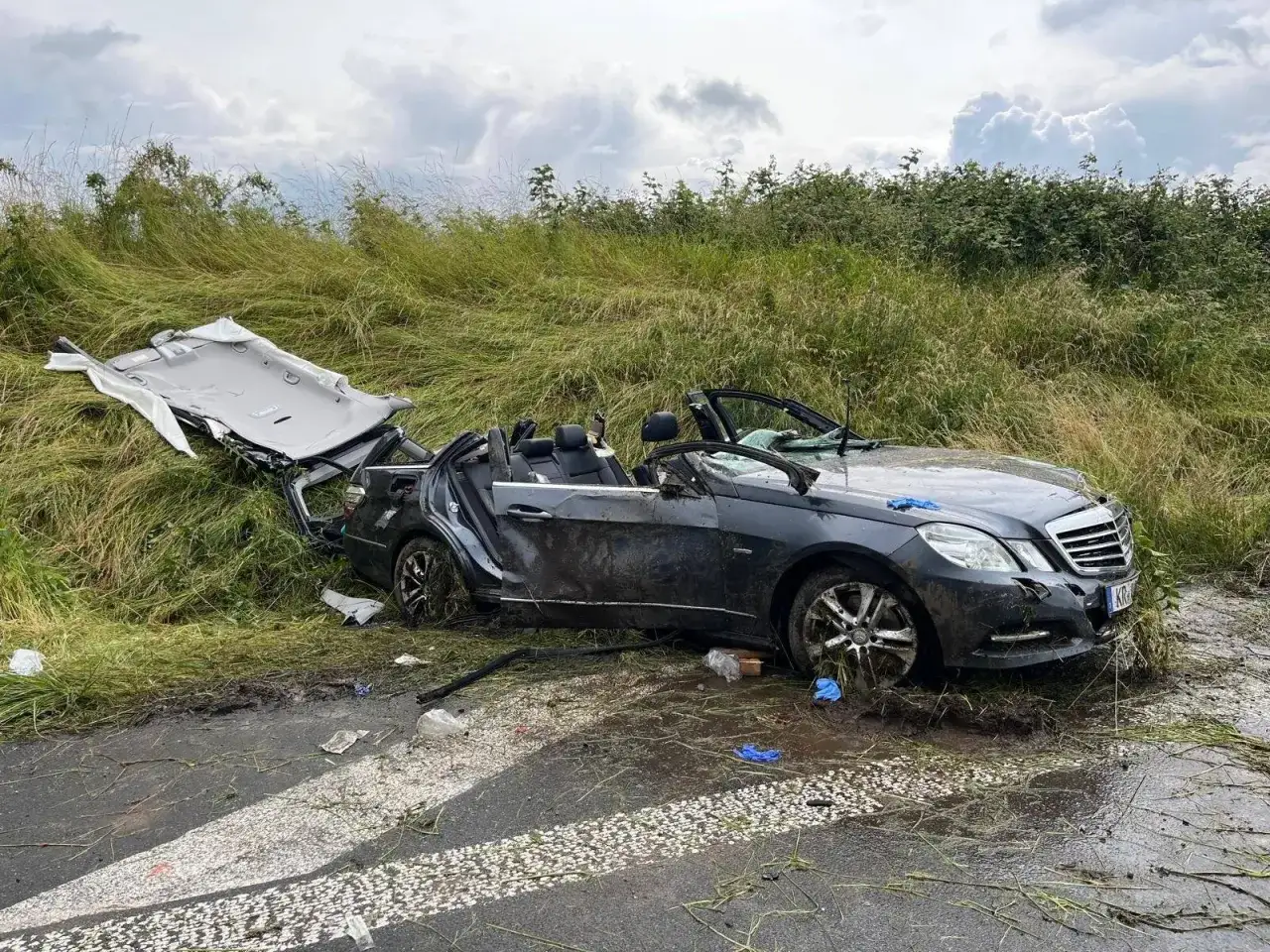 Ein dunkler Mercedes liegt nach einem Unfall schwer besch&auml;digt im Graben. Das Dach ist abgerissen, die T&uuml;ren offen. Tr&uuml;mmerteile und Schlamm bedecken die Fahrbahn.