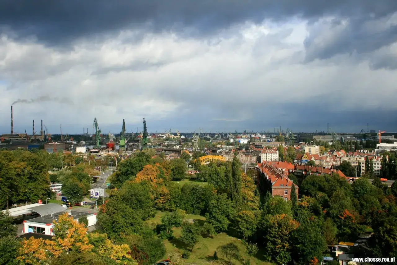Widok na portową część miasta z zielonymi dźwigami i kominami, na pierwszym planie jesienne drzewa. Góra Gradowa w oddali.