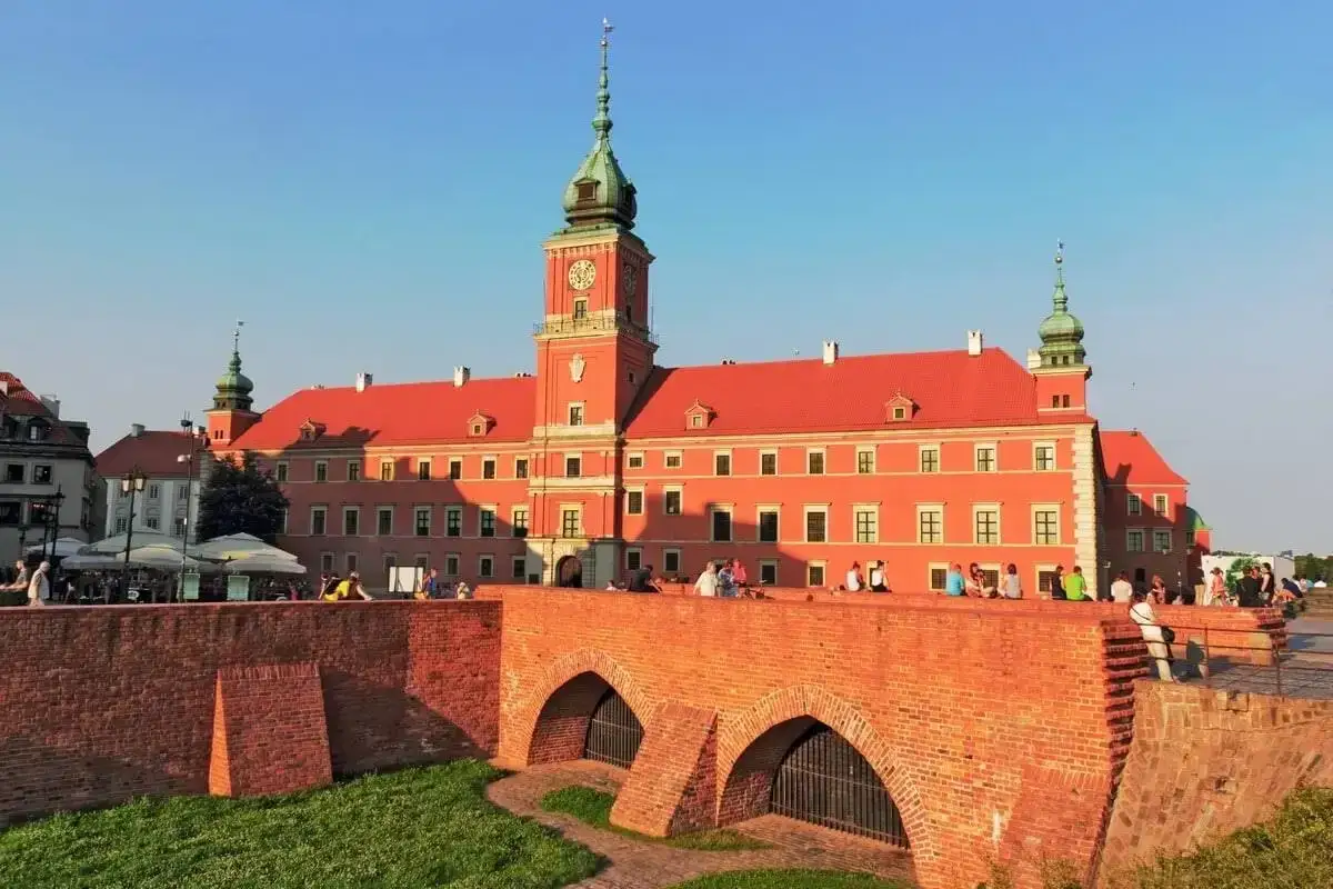Zamek Królewski, serce warszawskiego Starego Miasta, lśni w słońcu. Ludzie odpoczywają na ceglanym moście, podziwiając architekturę.