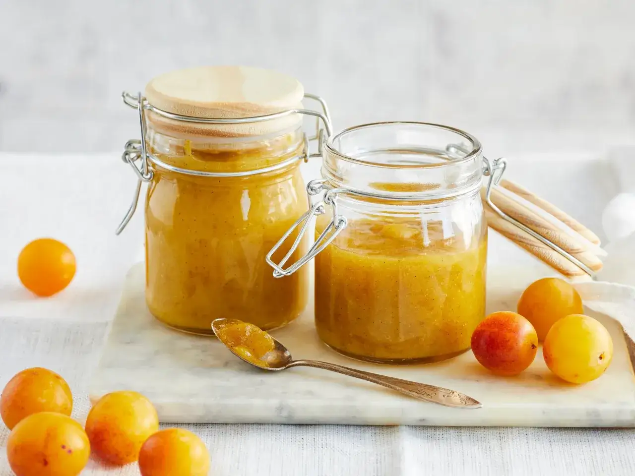 Zwei Gläser mit goldgelber Mirabellenmarmelade, daneben frische Mirabellen und ein Löffel voll Marmelade. Ein einfaches mirabellenmarmelade rezept.