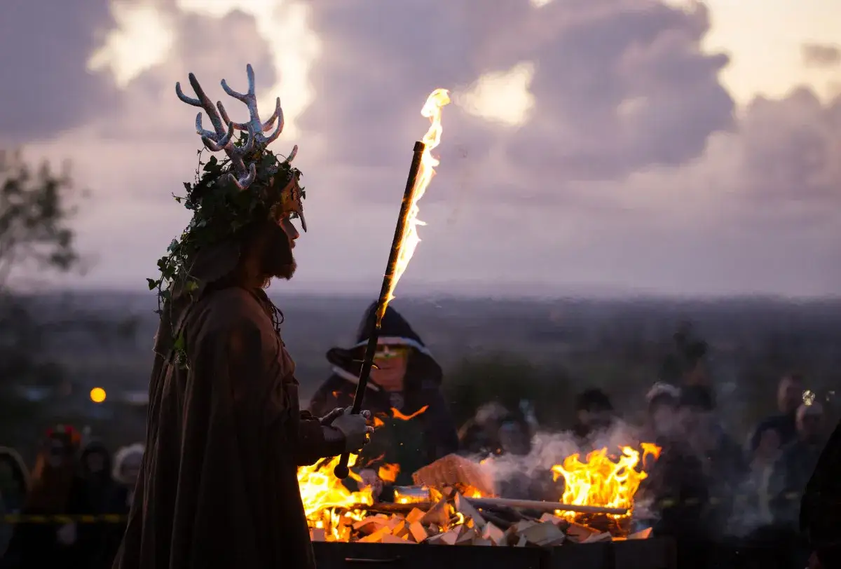 Mann mit Geweih-Kopfschmuck und Fackel bei einem Lagerfeuer. Dies könnte die wahre Bedeutung von Halloween symbolisieren: ein Fest des Lichts und der Gemeinschaft.
