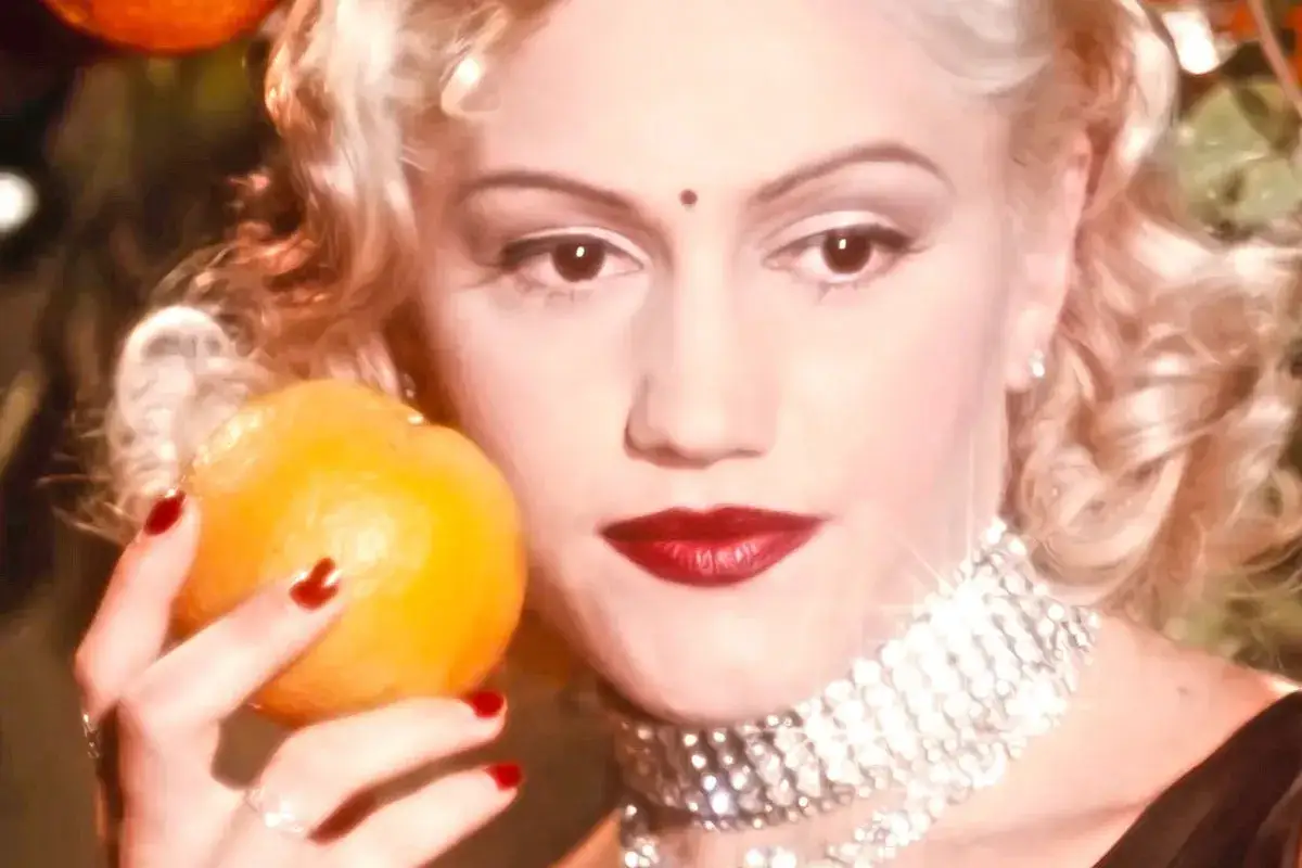 Gwen Stefani h&auml;lt eine Orange, tr&auml;gt roten Lippenstift und eine funkelnde Halskette.