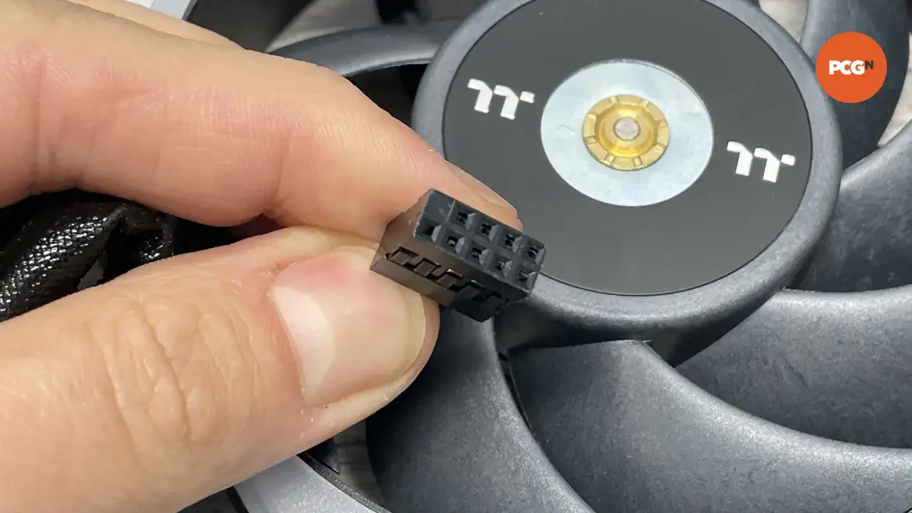 Ein Finger h&auml;lt einen schwarzen Stecker mit mehreren Pins, der f&uuml;r die RGB-Beleuchtung eines PC-L&uuml;fters gedacht ist.