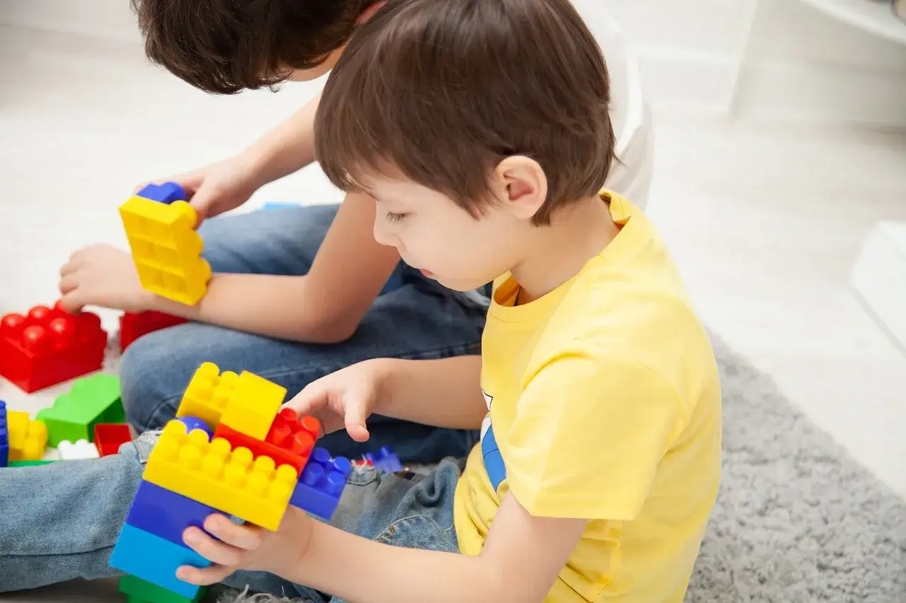 Bambini iperattivi 1 anno giocano con blocchi colorati. Un bimbo con maglietta gialla assembla una torre, mentre l'altro, pi&ugrave; grande, tiene un pezzo giallo.