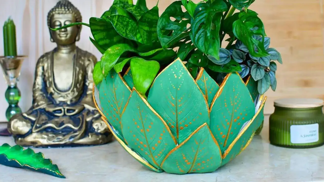 Zielona doniczka diy w kształcie liści, ozdobiona złotymi detalami, otacza bujną roślinność. Obok siedzi złoty Budda.
