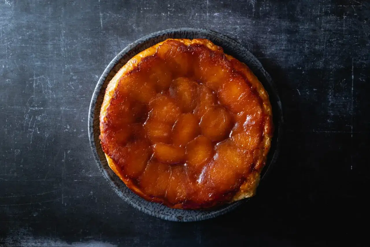 En gyllene, karamelliserad tarte tatin med äppelskivor på en mörk bakgrund.