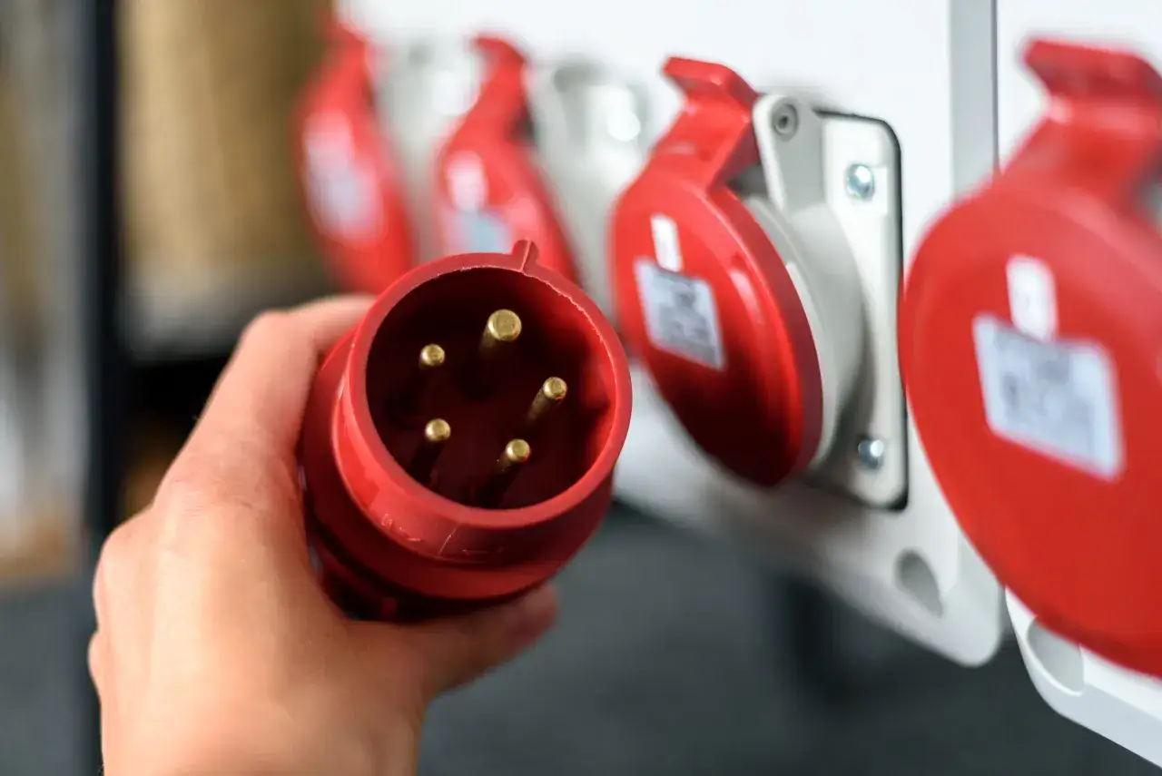 Dłoń trzyma wtyczkę elektryczną. W tle czerwone gniazda, jak w hurtowni art. elektrycznych Kowal K.