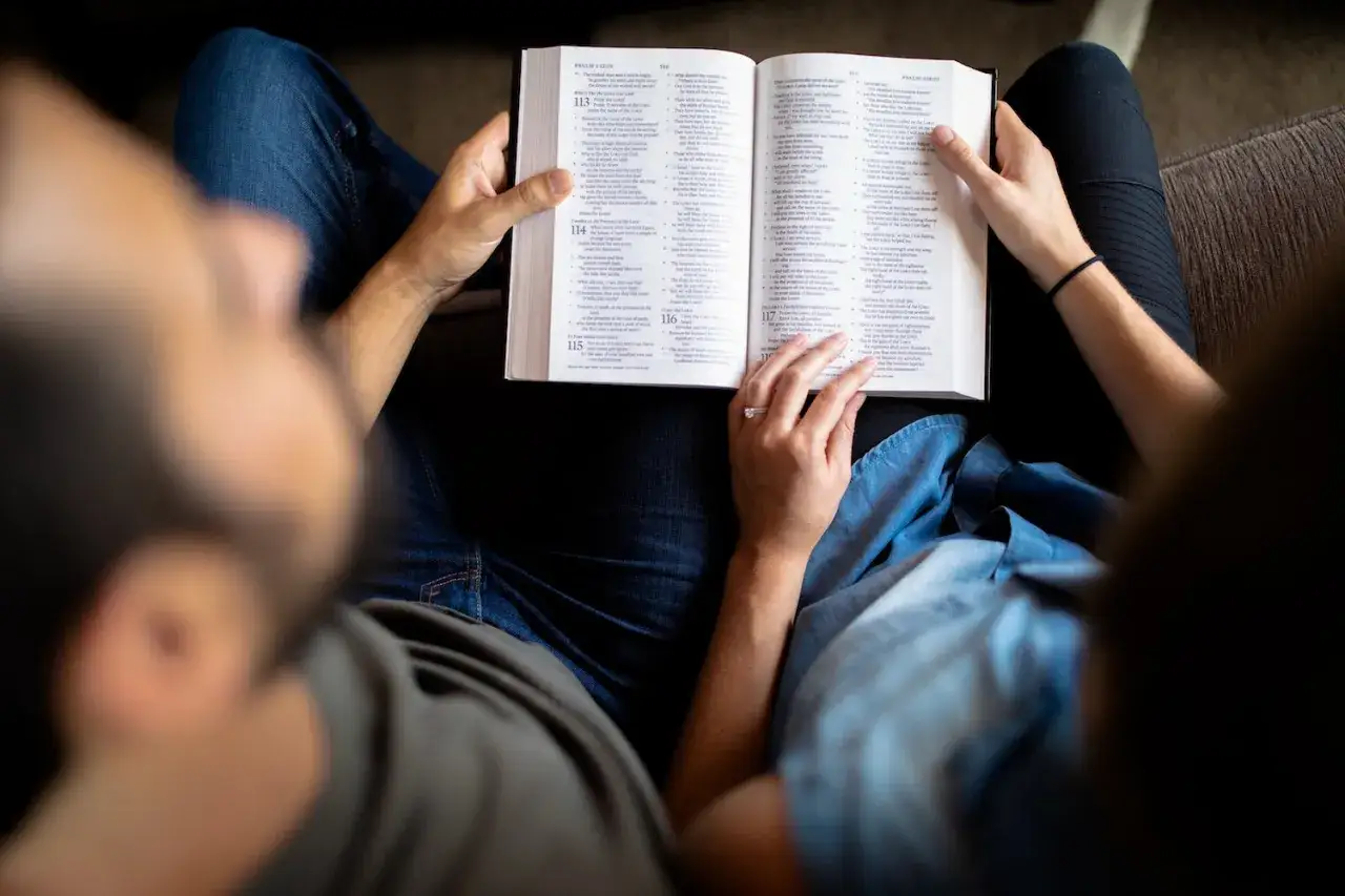 A couple engaged in a couples bible study, reading from an open Bible together on a couch.