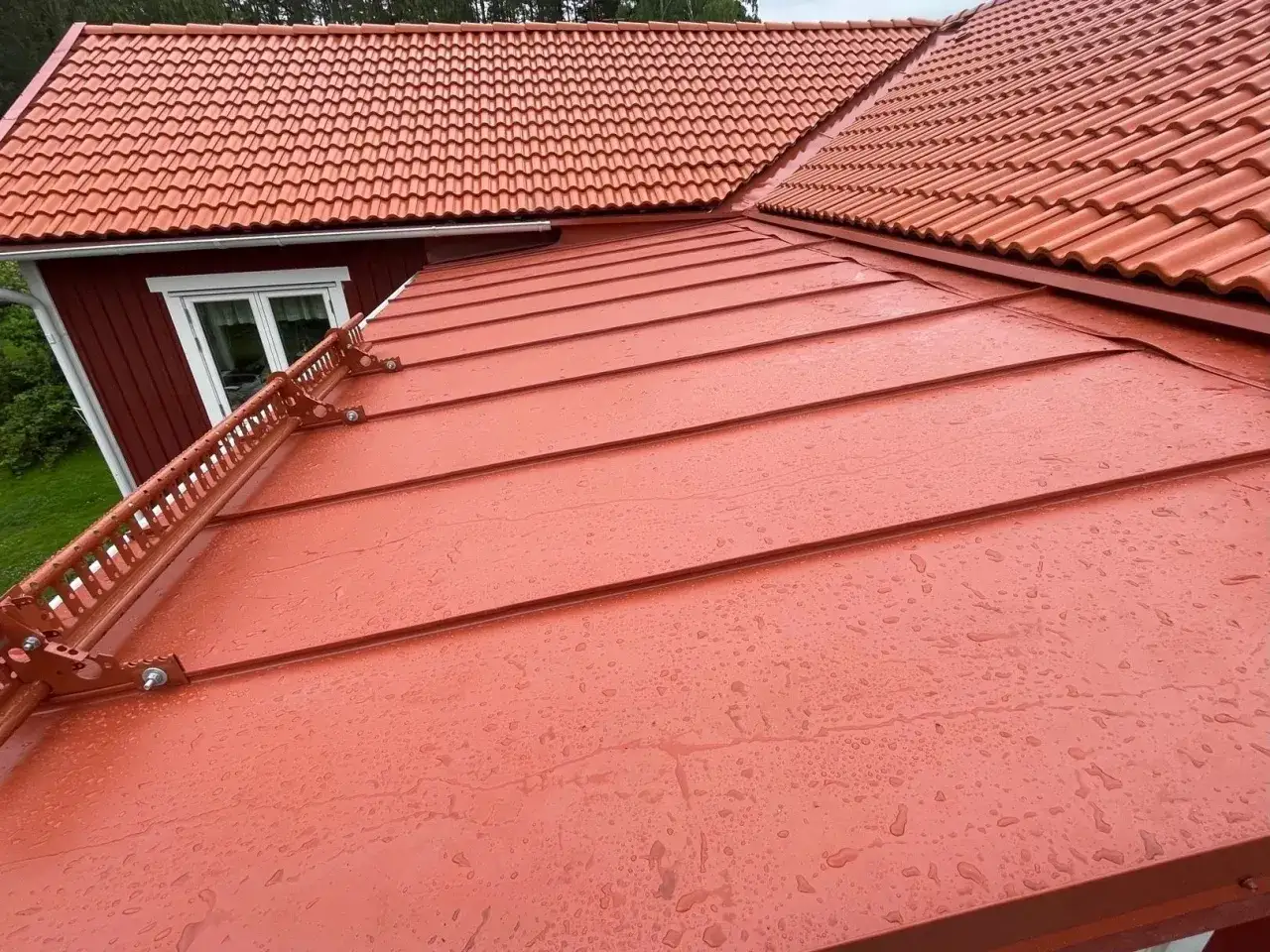 Ett r&ouml;tt, bandat pl&aring;ttak med vattendroppar. Till v&auml;nster syns en del av ett r&ouml;tt tr&auml;hus med ett tegeltak.