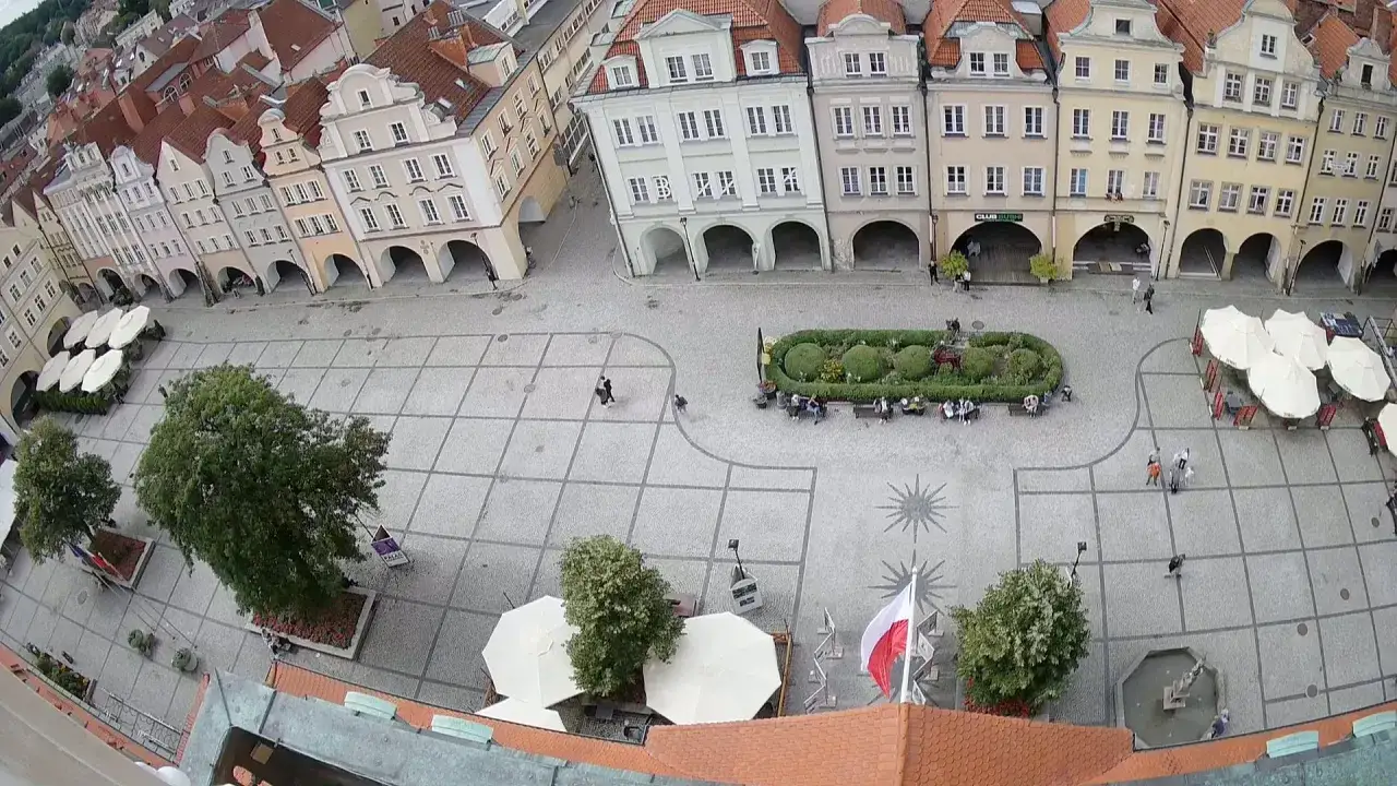 Urokliwy rynek w Jeleniej G&oacute;rze z kolorowymi kamieniczkami, zieloną wyspą i ludźmi spacerującymi po bruku.