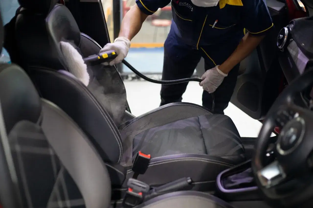 Persona con guantes usa vapor para limpiar tapicer&iacute;a coche. El vapor elimina suciedad y renueva asientos.
