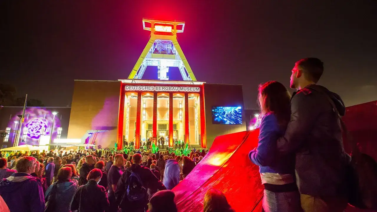 Ein Paar genießt die **Extraschicht Bochum** vor dem beleuchteten Bergbau-Museum. Eine Menschenmenge feiert unter dem Förderturm.
