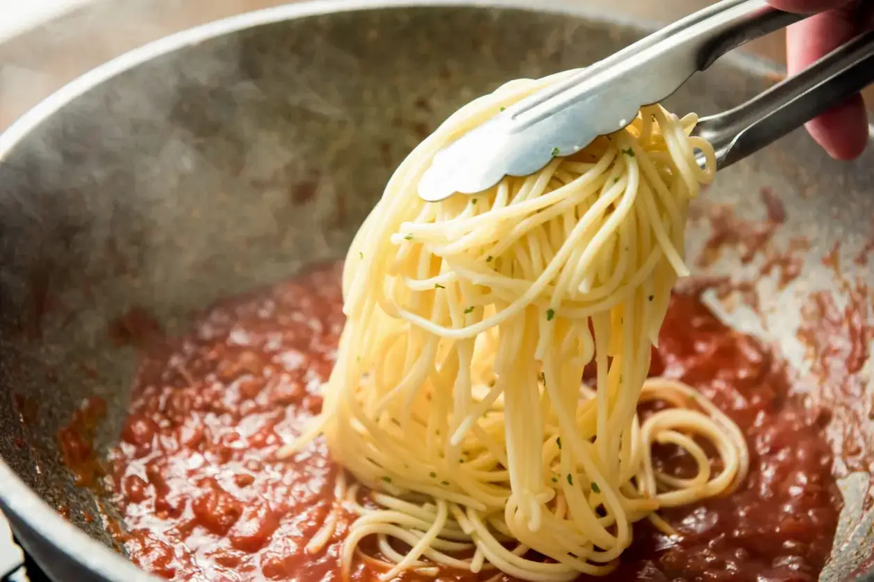 Szczypce podnoszą porcję spaghetti z sosu pomidorowego w patelni. Czy to bucatini czy spaghetti?