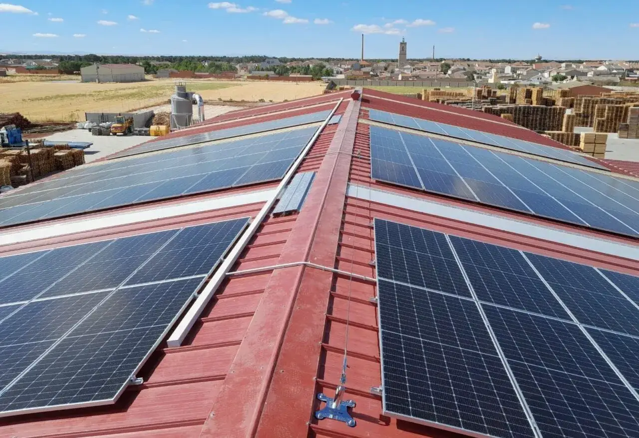 Amplia cubierta fotovoltaica sobre tejado rojo, captando energ&iacute;a solar. Al fondo, un pueblo y campos.