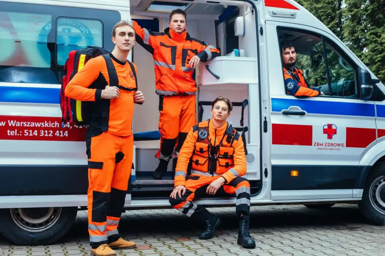 Trzech młodych ratowników medycznych w pomarańczowych strojach stoi obok karetki. Jeden z nich ma plecak ratowniczy.