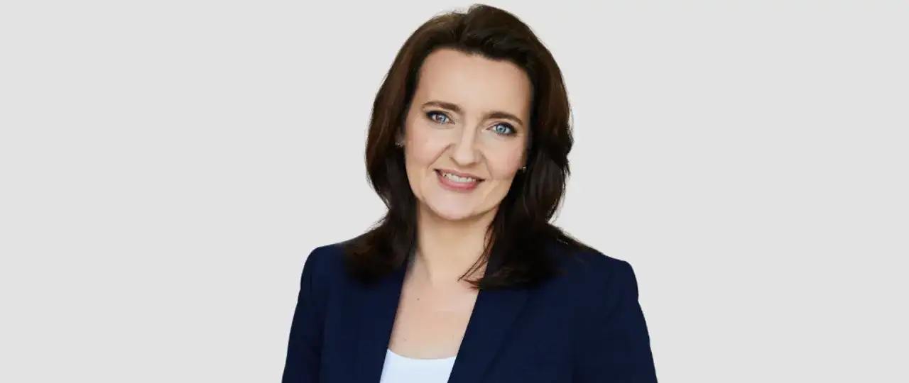 Uśmiechnięta Marzena Okła-Drewnowicz w granatowej marynarce, z niebieskimi oczami i brązowymi włosami.