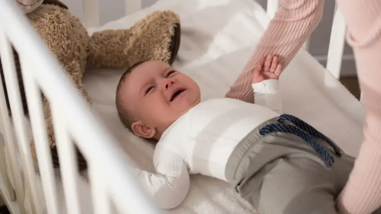 Beb&eacute; llorando en la cuna, con un peluche. Una mano lo sujeta, quiz&aacute;s aplicando el **m&eacute;todo dejar llorar al beb&eacute;**.