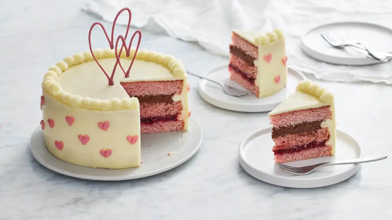 En rosa tårta med vit frosting, dekorerad med hjärtan. Skivor visar lager av rosa tårtbotten, choklad och bär. Perfekt för enkla tårtfyllningar.