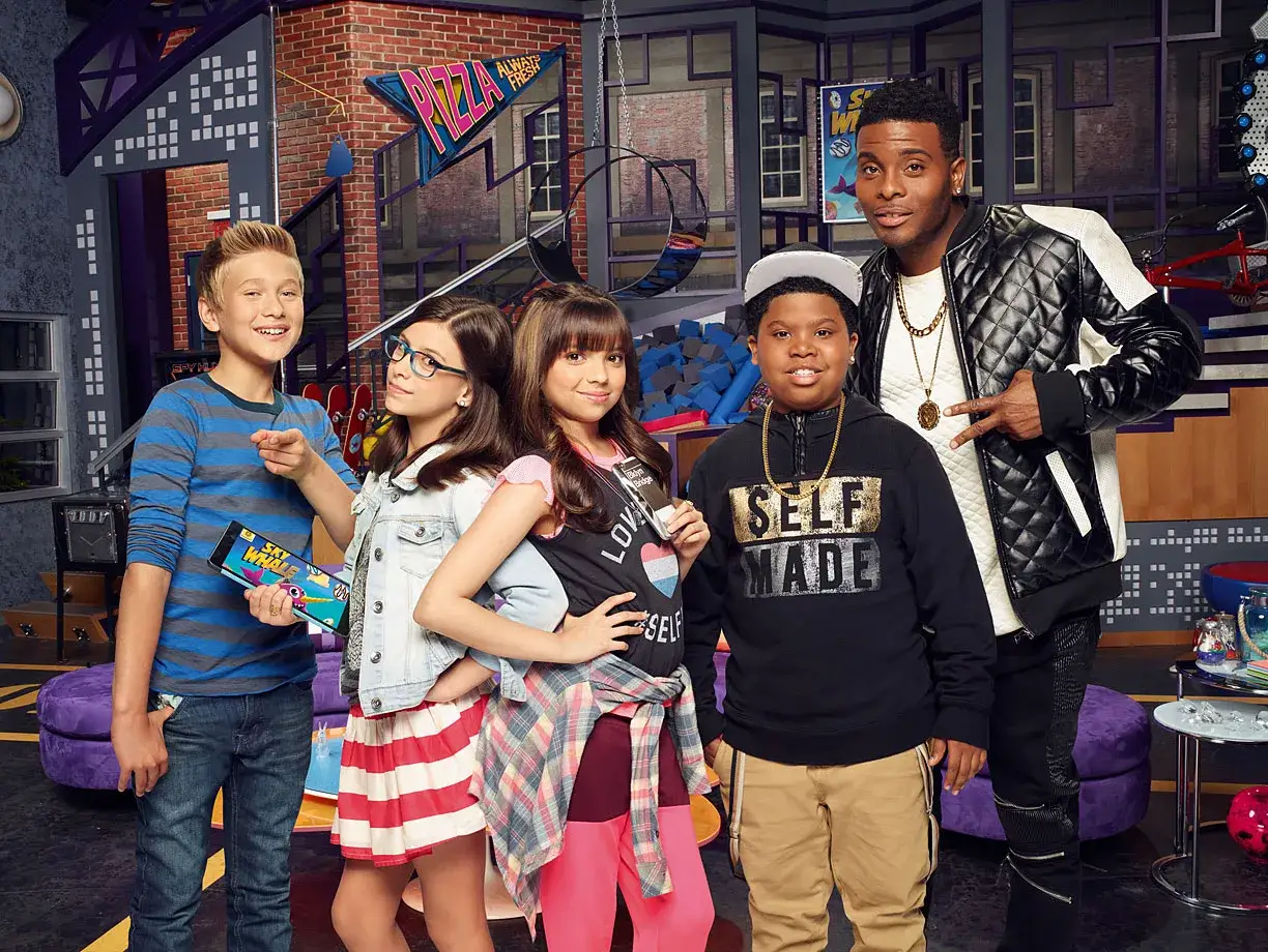 Grupa młodych twórców z Game Shakers, jak wydać grę? - w tym chłopiec z tabletem, dziewczynki w kolorowych strojach i dwaj starsi mężczyźni.