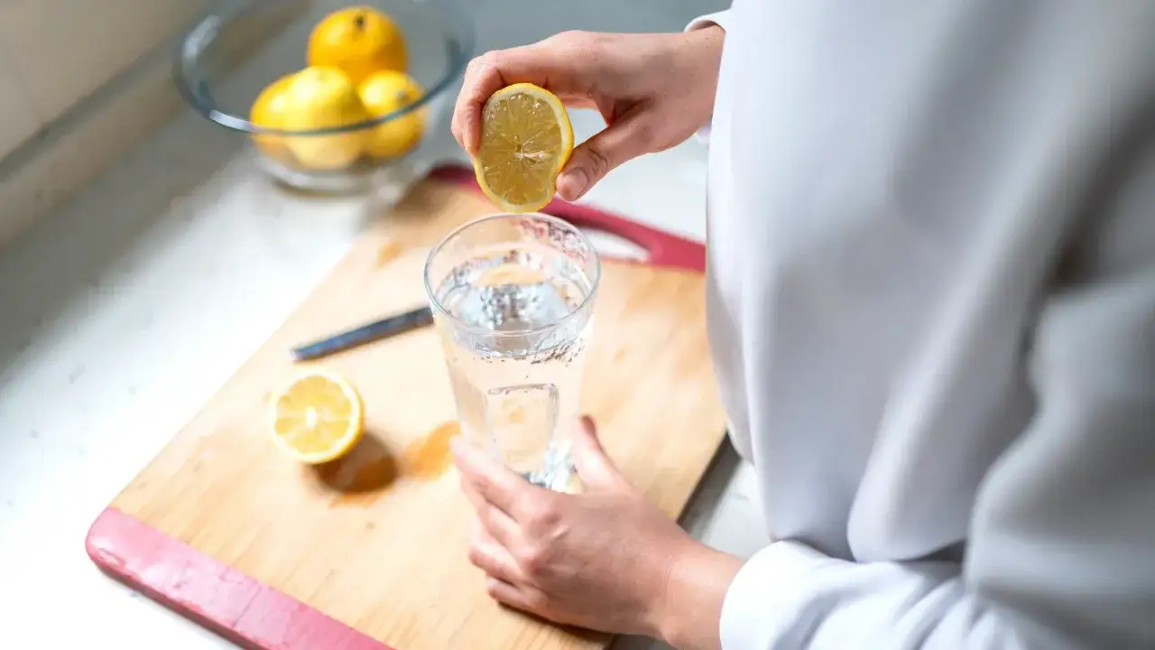Eine Hand drückt eine Zitronenhälfte über ein Glas Wasser. Ein gesunder Start in den Tag mit Wasser mit Zitrone morgens.