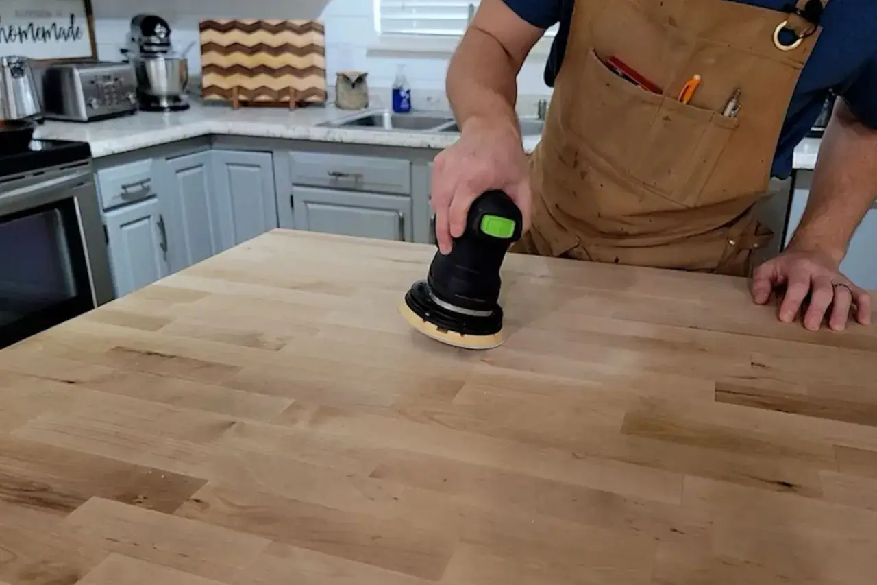 Un artisan utilise une ponceuse orbitale pour poncer un plan de travail en bois. La surface en bois est lisse et prête pour la finition.