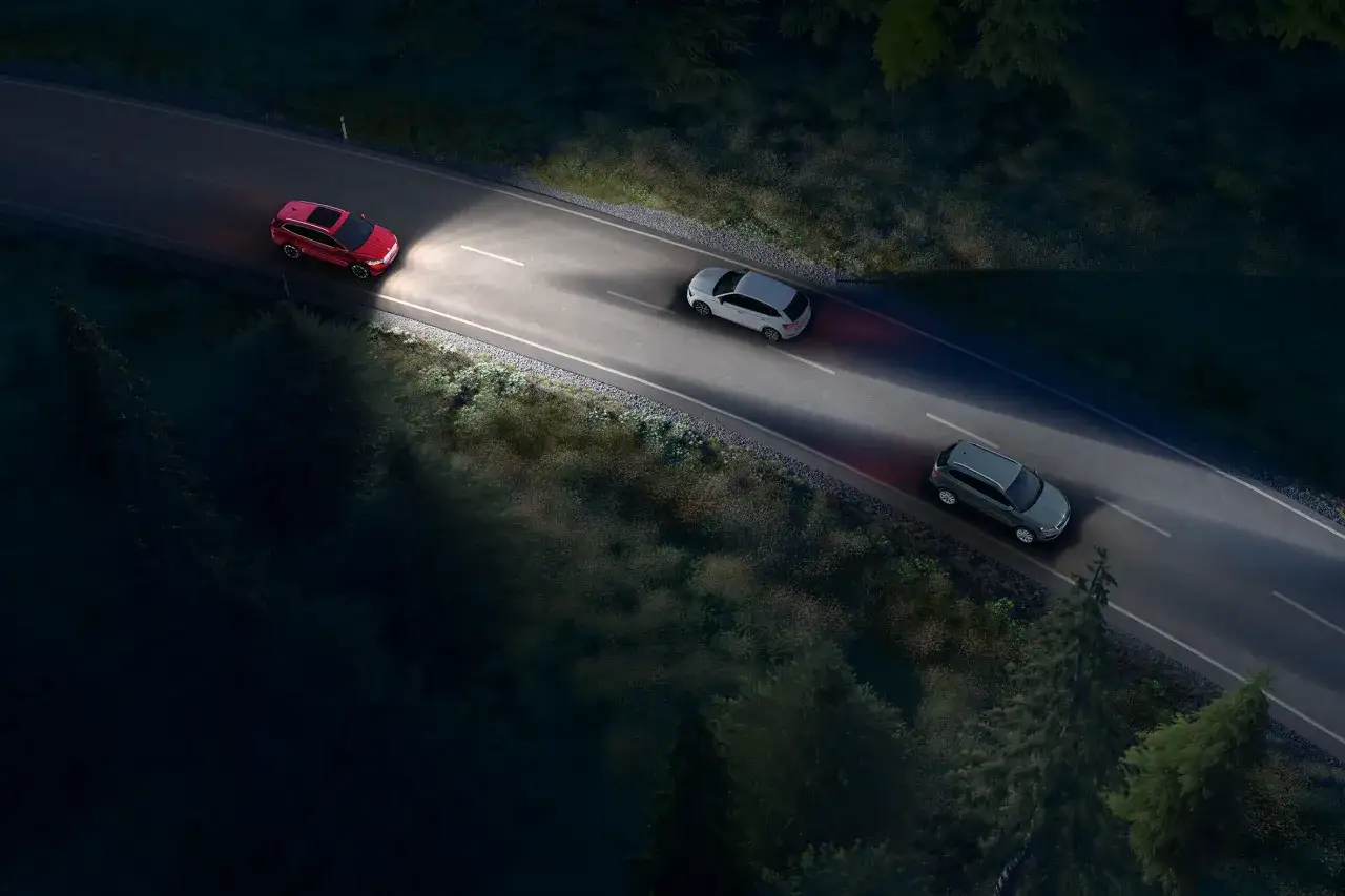 Trzy samochody jadą nocą po krętej drodze. Czerwony SUV z przodu używa świateł mijania, oświetlając drogę.