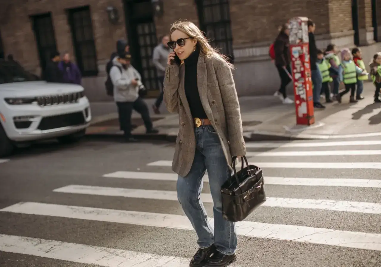 En kvinna i rutig kavaj, jeans och polotröja korsar gatan. En perfekt dagens outfit för en stadspromenad.