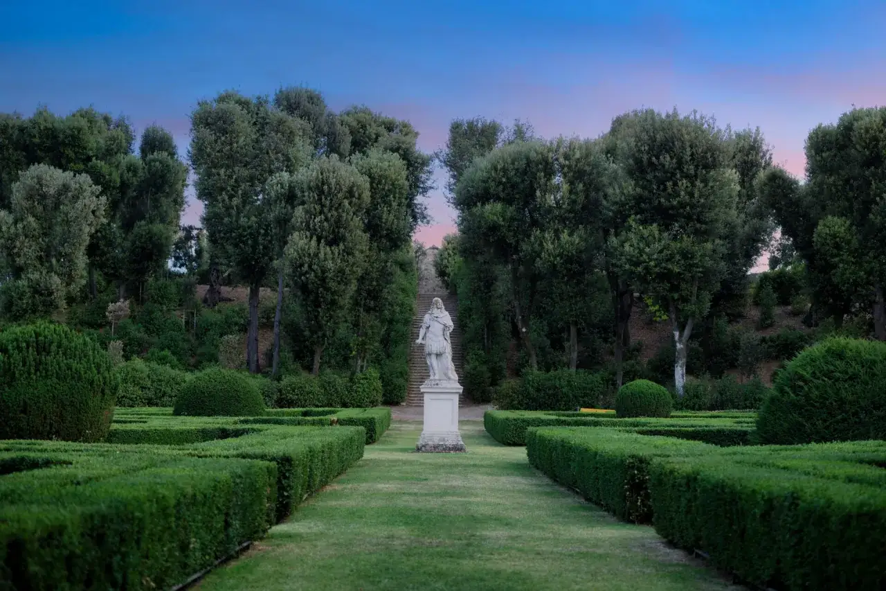 Statua al centro dei Horti Leonini, con siepi geometriche e alberi rigogliosi sotto un cielo crepuscolare.
