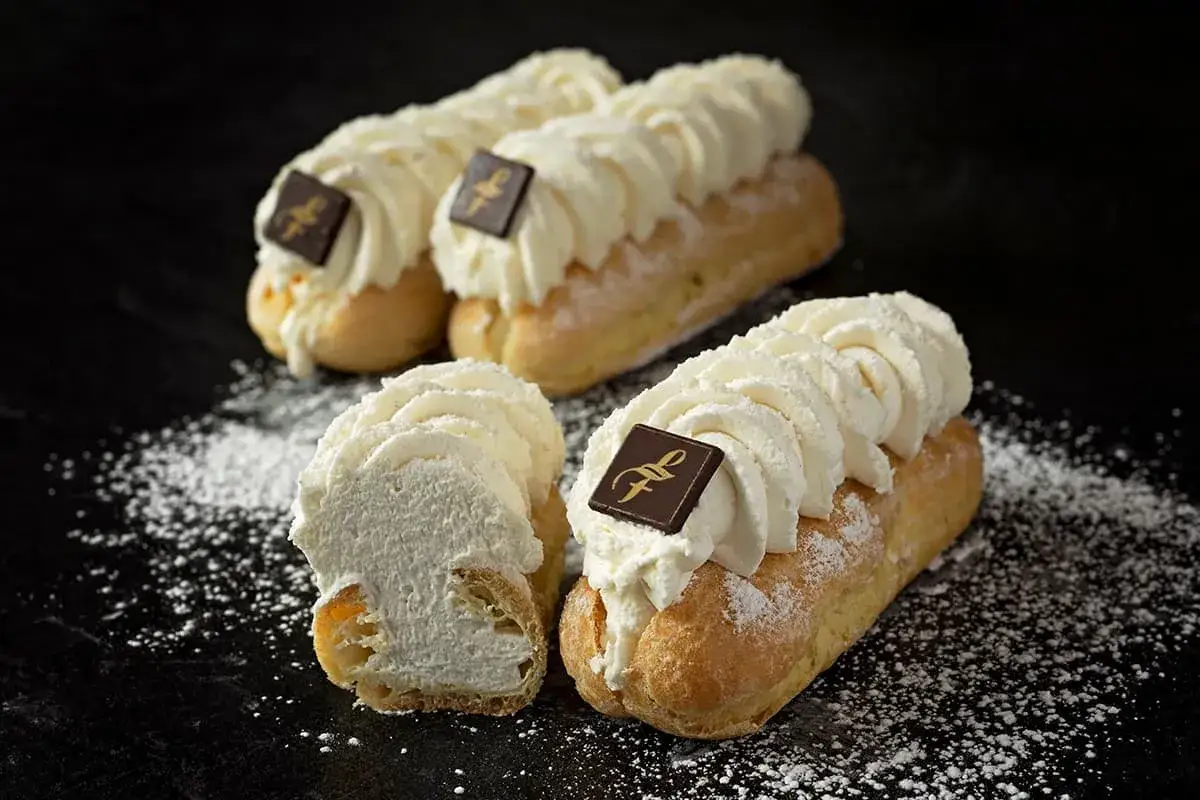 Trois éclairs chantilly généreusement garnis de crème fouettée, ornés d'un carré de chocolat, saupoudrés de sucre glace.