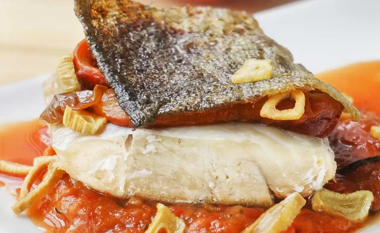 Bacalao a la riojana con piel crujiente, pimientos rojos y ajo frito, servido sobre una cama de salsa de tomate.