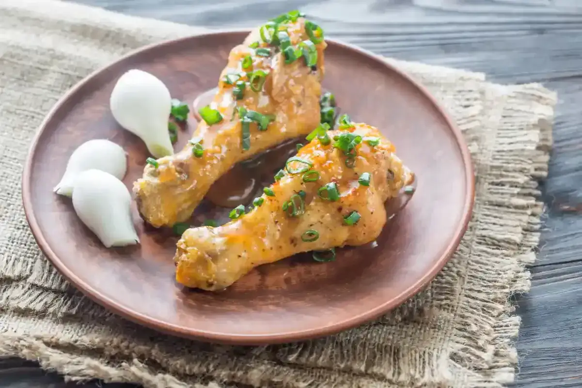 Deux pilons de poulet app&eacute;tissants, napp&eacute;s d'une sauce onctueuse et parsem&eacute;s de ciboule fra&icirc;che, accompagn&eacute;s de petits oignons blancs sur une assiette en terre cuite.