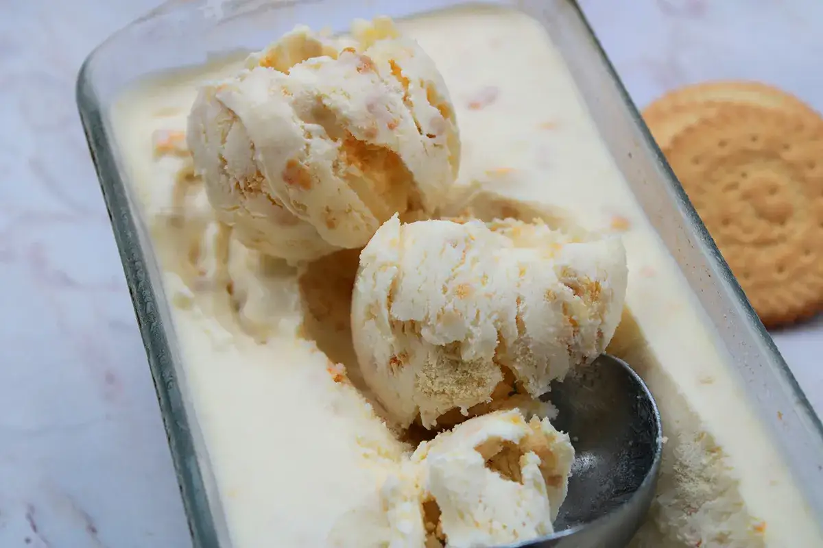 Pyszne lody ciasteczkowe w szklanym pojemniku, z widocznymi kawałkami ciastek i gałkami lod&oacute;w. Obok leży okrągłe ciastko.